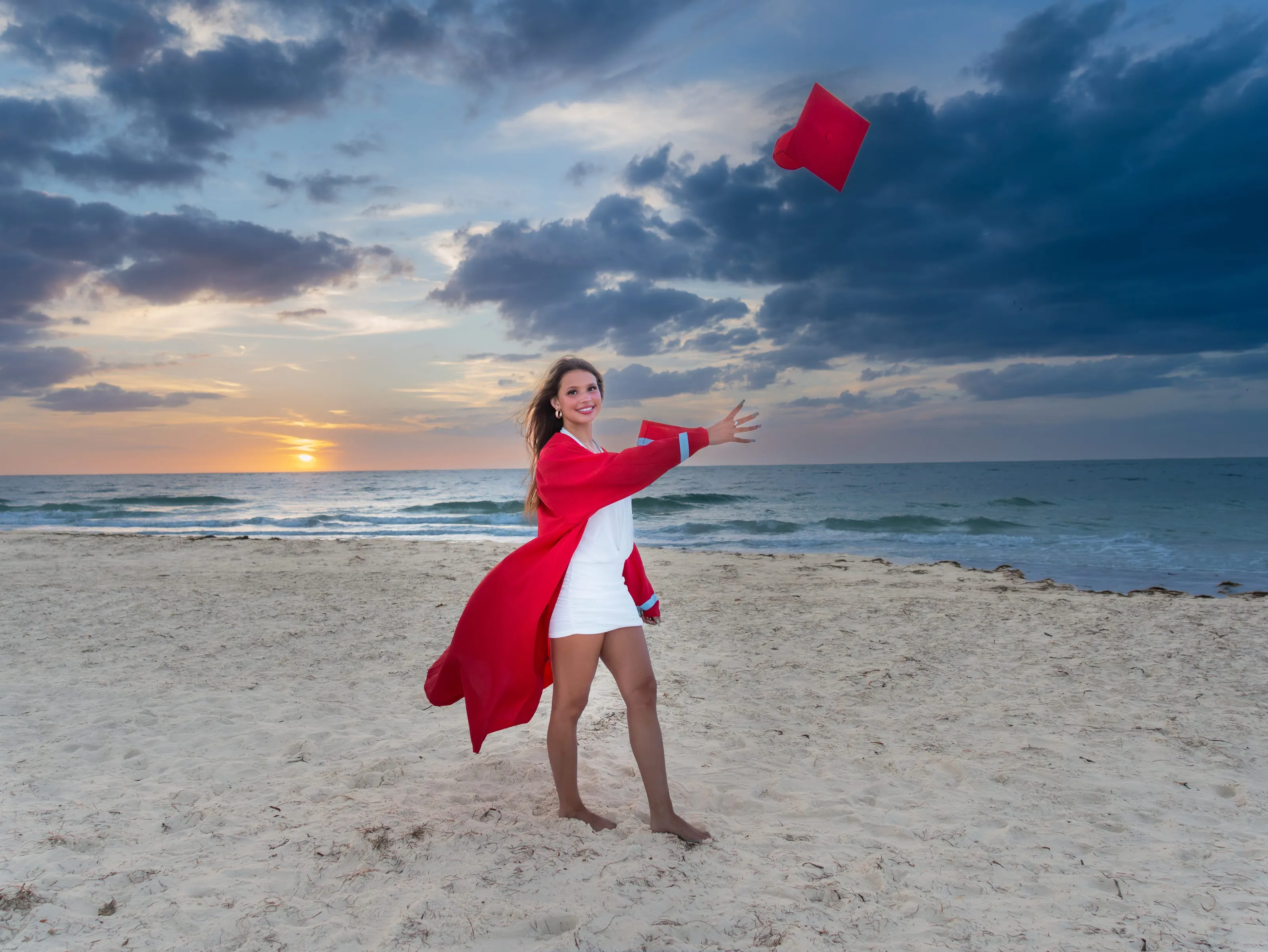 High school senior in a red graduation gown tossing her cap on the beach at sunset