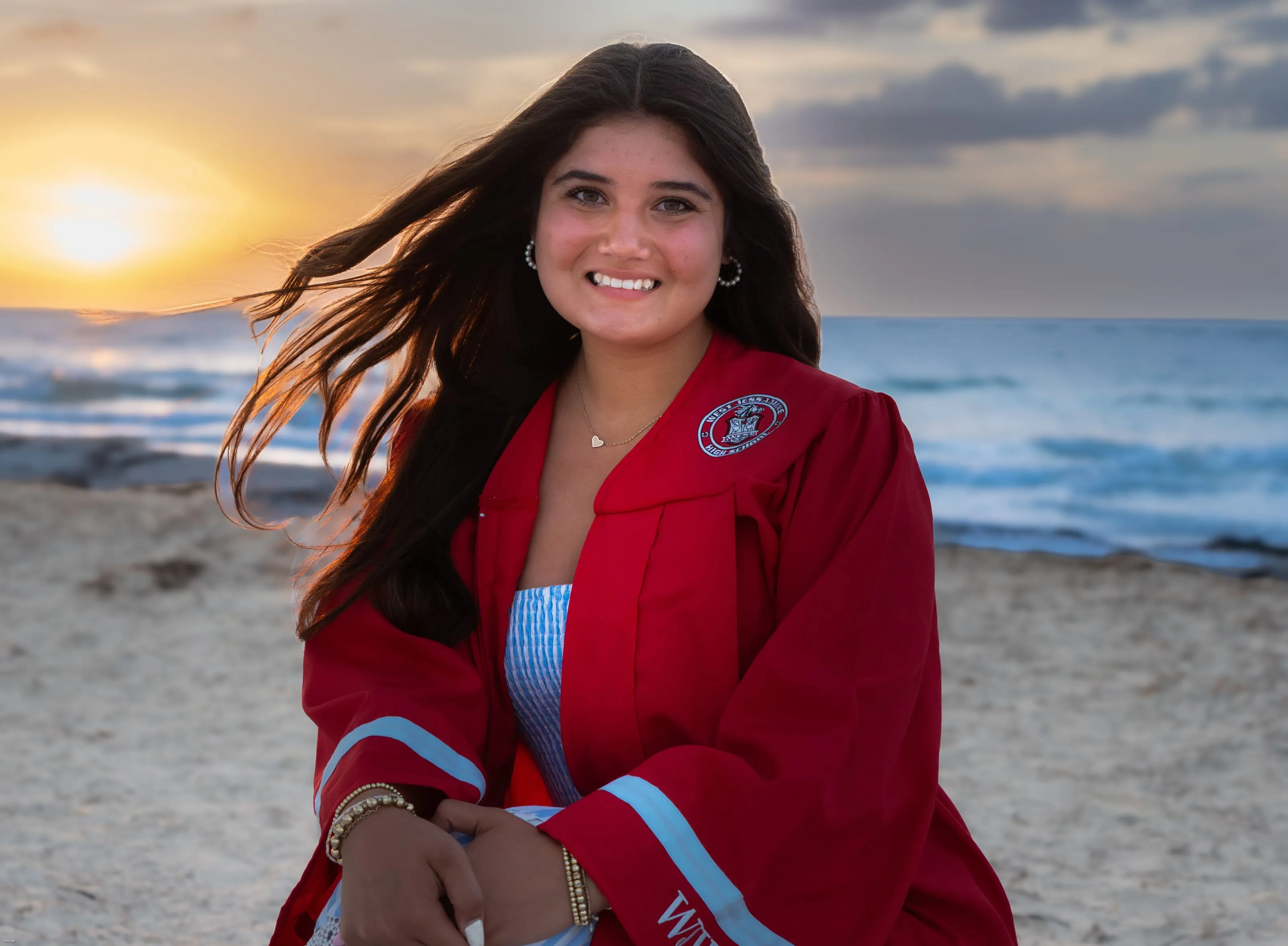 Smiling senior in a red graduation gown on the beach at sunset