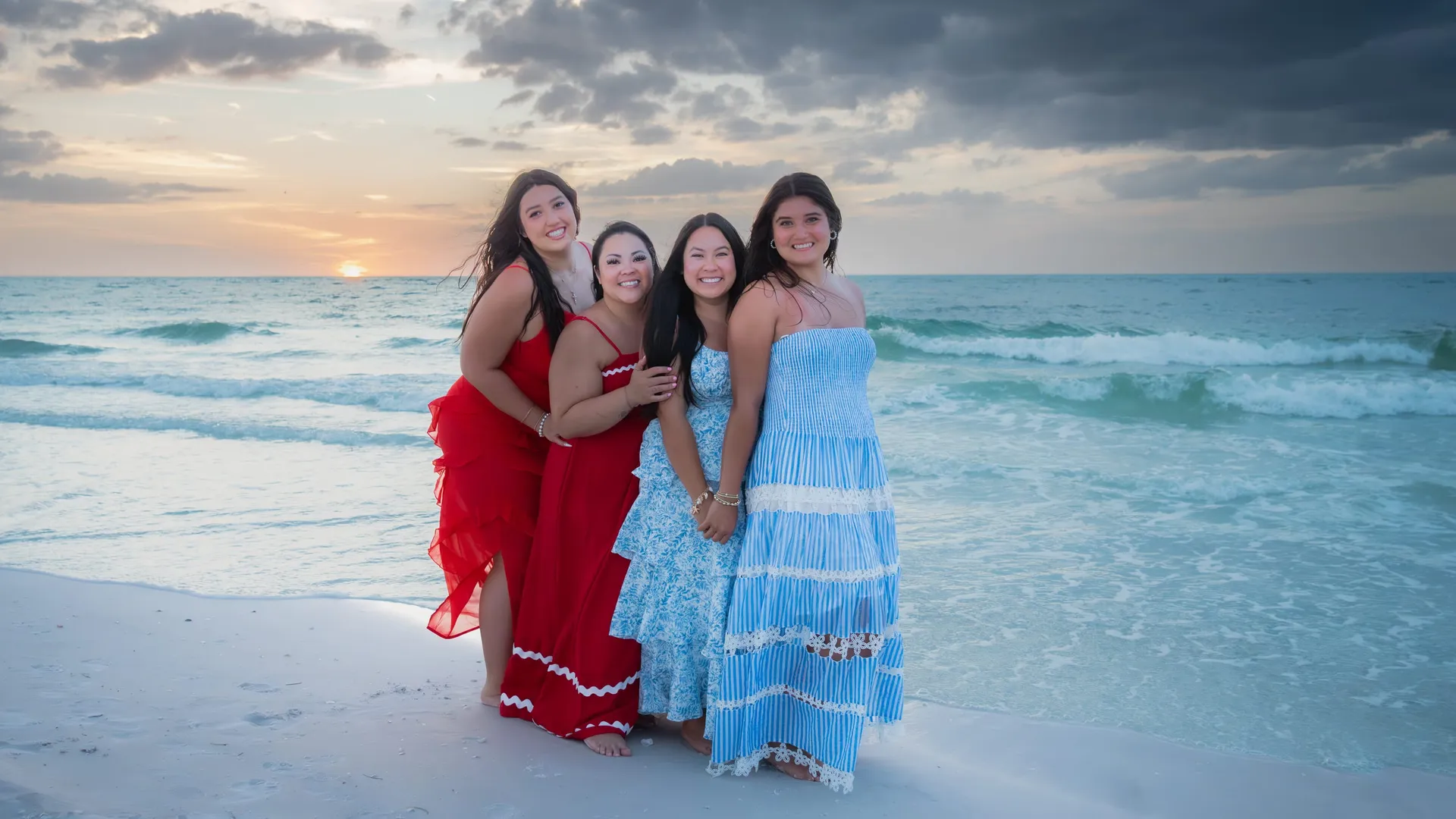 Four women in red, white, and blue dresses at the water’s edge at sunset